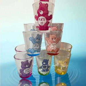 Könnte beinhalten: Eine Ansammlung klarer Schnapsgläser mit bunten Cartoon-Bären-Designs. Einige Gläser zeigen Texte wie "FUCK IT", "You're A Cunt" und "Smile Bitch". Die Gläser sind auf einer reflektierenden Oberfläche gestapelt.