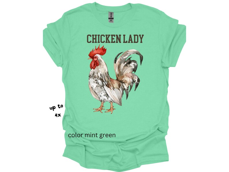May include: Mint green t-shirt with the text "CHICKEN LADY" above a watercolor illustration of a rooster. The rooster has red, brown, white, and gray feathers. The shirt is available in sizes up to 4X.