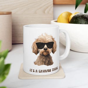 May include: White ceramic mug featuring a brown Cavapoo dog wearing black sunglasses. The mug has the phrase "IT'S A CAVAPOO THING" printed in black. The mug is on a beige coaster.