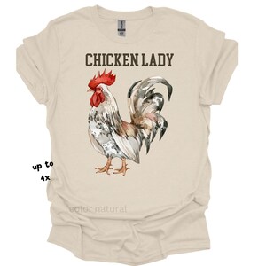 May include: A cream-colored t-shirt featuring a watercolor illustration of a rooster with red comb and wattle, and gray and white feathers. The text "CHICKEN LADY" is printed above the rooster. The shirt is available in sizes up to 4X.