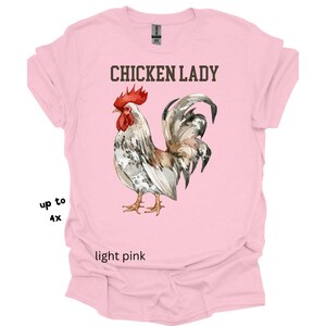 May include: Light pink t-shirt with the text "CHICKEN LADY" above a watercolor illustration of a rooster. The rooster has red, white, brown, and gray feathers. The shirt is available up to size 4X.