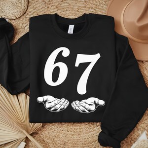 May include: Black sweatshirt with the number 67 in white, stylized font. Below the number are two white, open hands. The sweatshirt is displayed on a woven surface with a straw hat and a woven basket in the background.