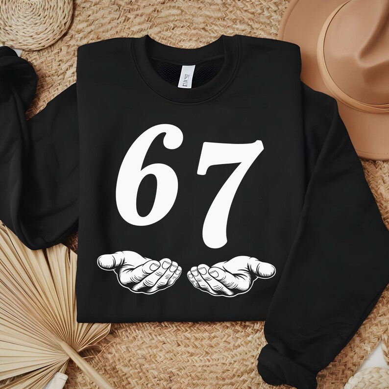 May include: Black sweatshirt with the number 67 in white, stylized font. Below the number are two white, open hands. The sweatshirt is displayed on a woven surface with a straw hat and a woven basket in the background.