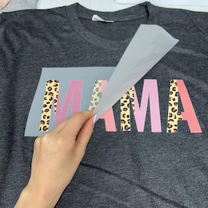 May include: Dark gray t-shirt with the word "MAMA" in pink and leopard print lettering. A hand is peeling away a clear transfer sheet, revealing the design. The shirt is made of a soft material.