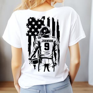 Pode incluir: Camiseta branca com uma ilustra&ccedil;&atilde;o a preto e branco de um jogador de futebol americano. O design inclui uma bandeira americana, o nome "JOHNSON" e o n&uacute;mero "9". O jogador est&aacute; segurando uma bola de futebol.