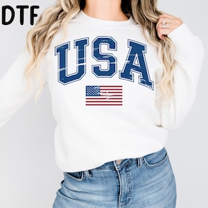 May include: White sweatshirt with the word "USA" in distressed blue lettering and a small American flag graphic. The sweatshirt is a crew neck style and is made of a soft material.