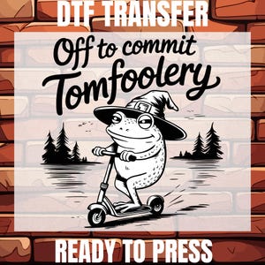 May include: A black and white illustration of a frog wearing a witch's hat and riding a scooter. The frog is riding through a forest and the text "Off to commit tomfoolery" is above the frog. The text "DTG Transfer Ready to Press" is below the frog.