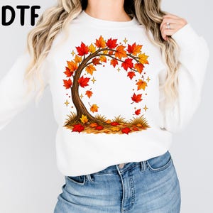 May include: A white long-sleeve sweatshirt featuring a central graphic of a brown tree with autumn leaves in shades of red, orange, and yellow. The leaves are depicted falling and scattered on the ground around the base of the tree trunk.