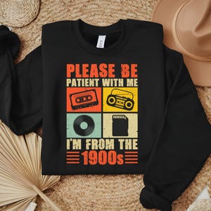 May include: Black sweatshirt with the text "PLEASE BE PATIENT WITH ME I'M FROM THE 1900s" and retro graphics of a cassette tape, boombox, vinyl record, and SD card. The text and graphics are in orange, red, yellow, and teal.