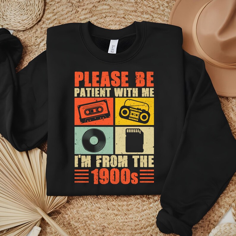 May include: Black sweatshirt with the text "PLEASE BE PATIENT WITH ME I'M FROM THE 1900s" and retro graphics of a cassette tape, boombox, vinyl record, and SD card. The text and graphics are in orange, red, yellow, and teal.