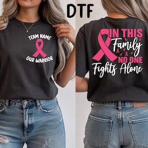 May include: A dark gray t-shirt featuring a pink awareness ribbon on the front left chest with the text Team Name Our Warrior. The back displays a large pink ribbon and the text In This Family No One Fights Alone in pink and white lettering.