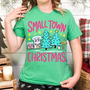 May include: Green t-shirt with the words "SMALL TOWN CHRISTMAS" in pink. The graphic includes cartoon houses, Christmas trees, stars, and a sign with the number 38864. The shirt is a festive holiday design.