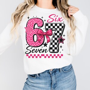 Puede incluir: Sudadera blanca con un gr&aacute;fico rosa y negro que dice "Six Seven". El n&uacute;mero 6 es rosa con un estampado de leopardo, y el n&uacute;mero 7 es un patr&oacute;n de cuadros blancos y negros. Una cinta rosa est&aacute; en el centro.