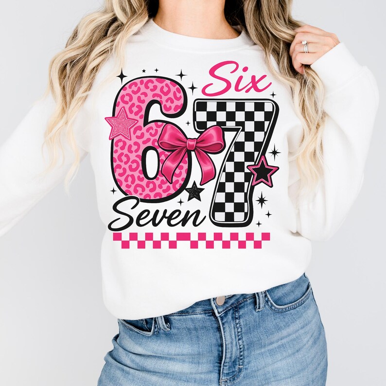 Puede incluir: Sudadera blanca con un gr&aacute;fico rosa y negro que dice "Six Seven". El n&uacute;mero 6 es rosa con un estampado de leopardo, y el n&uacute;mero 7 es un patr&oacute;n de cuadros blancos y negros. Una cinta rosa est&aacute; en el centro.