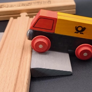 Pode incluir: Um comboio de brinquedo de madeira com cabine vermelha e secção de carga amarela, com rodas vermelhas. O comboio tem uma base preta e está posicionado num bloco cinzento, com carris de madeira no fundo. O comboio tem o logótipo Deutsche Post.