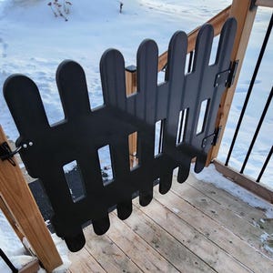 May include: Black plastic gate with a picket fence design, open on a wooden deck with snow on the ground.
