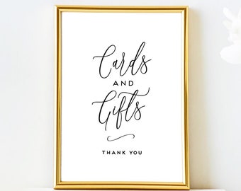 May include: A gold frame with a white background and black text that reads "Cards and Gifts Thank You". The frame is sitting on a stack of white books. There are white flowers in a clear vase to the right of the frame.