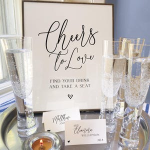 May include: A silver tray holds champagne flutes filled with bubbly liquid, a lit candle, and place cards. A framed sign reads "Cheers to Love" with additional text. The scene suggests a wedding or celebration.