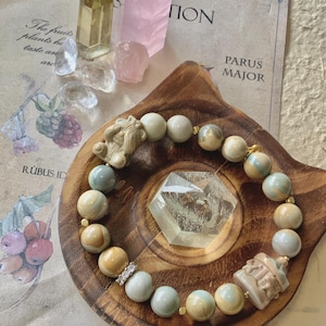 May include: A light blue and beige gemstone bracelet with gold accents. The bracelet is on a wooden surface with a clear crystal and a small figurine. The text "PARUS MAJOR" is visible in the background.