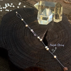 May include: A delicate bracelet featuring small pearls and amethyst beads strung on a purple string. The bracelet is displayed on a dark wooden surface, with the text "Purple String" and a few clear quartz crystals in the background.