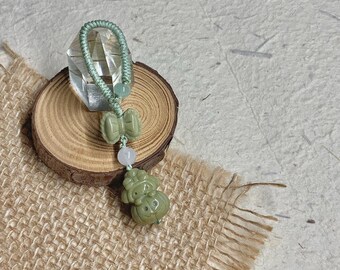 May include: A jade pendant with a green cord loop, featuring a carved gourd and other jade elements. The pendant is displayed on a small wooden slice and a textured tan fabric.