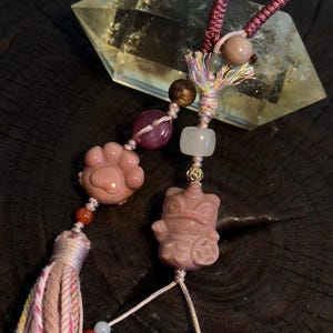 Puede incluir: Primer plano de un dije de piedra rosa con una huella de pata y una figura de rana. El dije está ensartado con un cordón rosa y adornado con cuentas y una borla. Un gran cristal transparente está en el fondo.