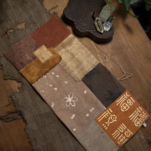 Puede incluir: Un tapiz textil rectangular con un diseño de patchwork de varios cuadrados de tela en tonos marrón, bronceado y óxido. La pieza incluye una sección con un patrón floral blanco y una sección con caracteres chinos blancos estilizados.