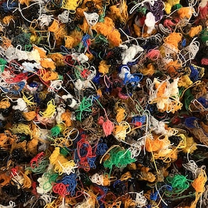 May include: A dense, chaotic collection of tangled plastic filaments in a vibrant array of colors including orange, yellow, blue, green, red, white, and black. The thin, extruded strands form a complex, knotted mass of overlapping loops.
