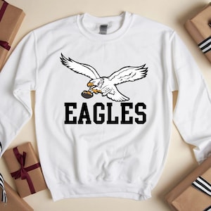 May include: White sweatshirt with a graphic of a soaring eagle holding a football, and the word "EAGLES" in black. The sweatshirt is surrounded by wrapped gifts with ribbons.