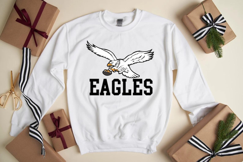 May include: White sweatshirt with a graphic of a soaring eagle holding a football, and the word "EAGLES" in black. The sweatshirt is surrounded by wrapped gifts with ribbons.