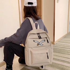 May include: Beige backpack with a front pocket and black zippers. The backpack features an embroidered eagle design on the front. The bag has adjustable shoulder straps and a top handle for carrying.