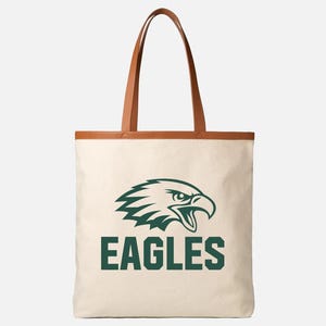 May include: Cream tote bag with brown leather handles and trim. The bag displays a green eagle head logo and the word "EAGLES" in green block letters. A stylish and practical accessory.