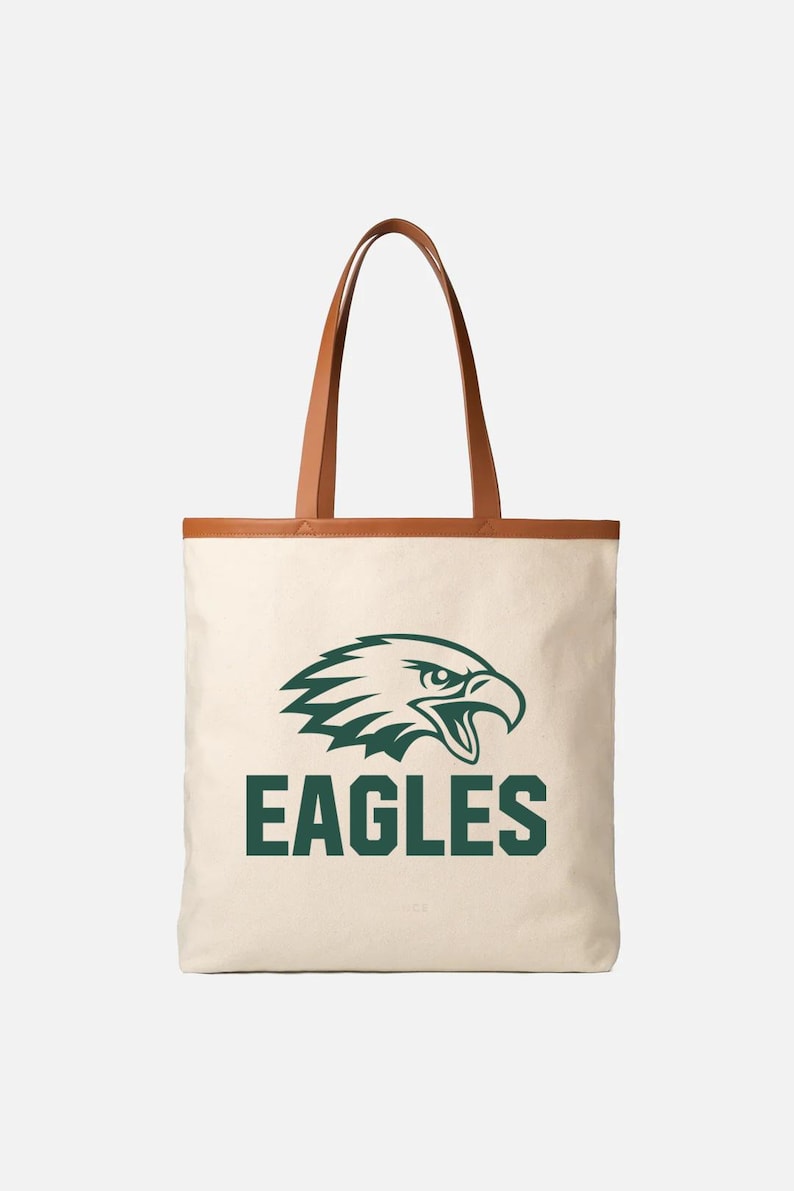 May include: Cream tote bag with brown leather handles and trim. The bag displays a green eagle head logo and the word "EAGLES" in green block letters. A stylish and practical accessory.