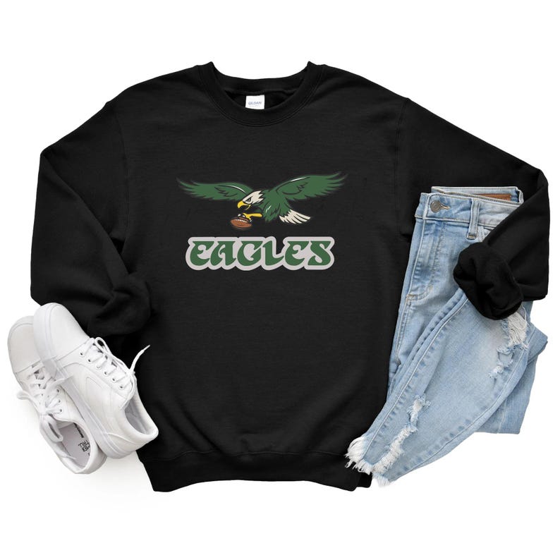 May include: Black sweatshirt with a green and white eagle graphic holding a football, and the word "EAGLES". The image includes light blue jeans and white trainers.