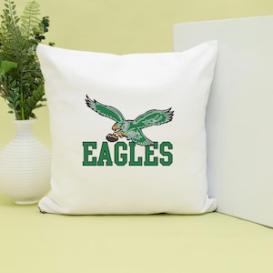May include: White square cushion featuring a green and white eagle graphic holding a football. The word "EAGLES" is printed in green block letters below the eagle. The cushion is a decorative item.
