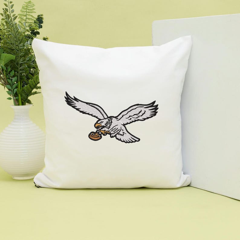 May include: White square pillow featuring an embroidered eagle in flight, clutching a football. The eagle is detailed in white and black, with a brown football. The pillow is a decorative item.