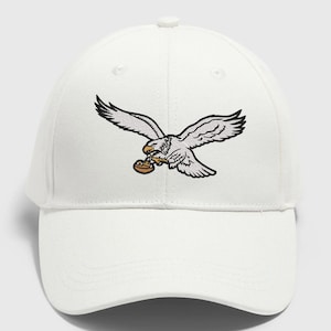 May include: White baseball cap featuring an embroidered eagle in flight, clutching a football. The eagle is depicted with black and white details, and the football is brown. The cap has a curved brim and a structured crown.