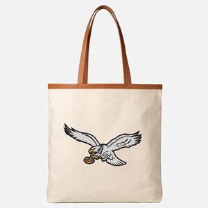 May include: Cream-colored tote bag with a brown leather trim and handles. The bag features an embroidered eagle design in gray, black, and brown. The eagle is depicted in flight, holding an object in its talons.