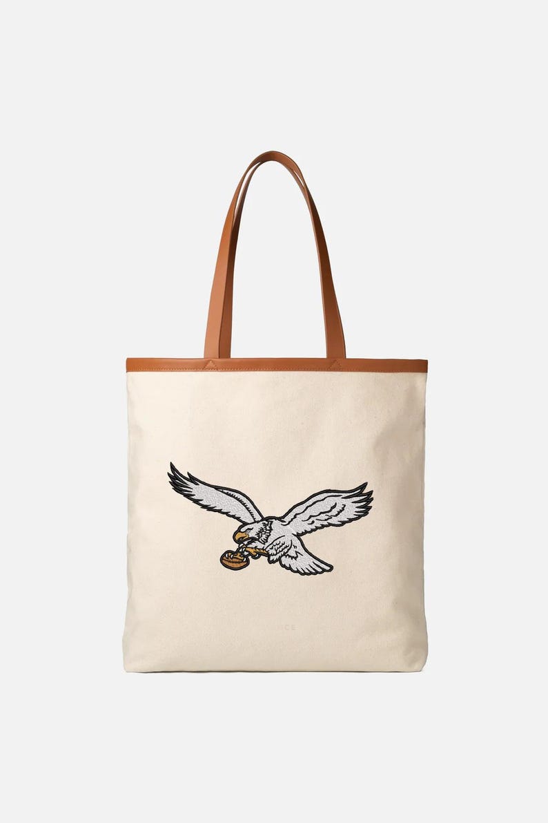 May include: Cream-colored tote bag with a brown leather trim and handles. The bag features an embroidered eagle design in gray, black, and brown. The eagle is depicted in flight, holding an object in its talons.