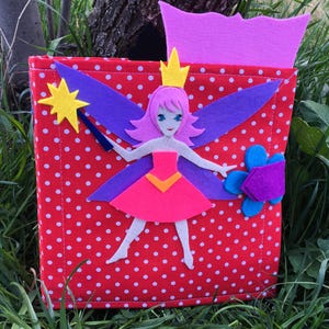 May include: A red felt book with white polka dots features a felt fairy with purple wings, pink hair, a yellow crown, and a pink dress. The fairy is holding a yellow star wand. The book also has a purple felt flower with a blue center.