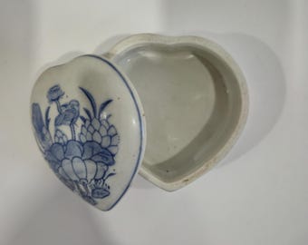 May include: Heart-shaped ceramic trinket box with a lid. The lid features a blue and white floral design. The box is open, revealing the interior. The box is on a white surface.