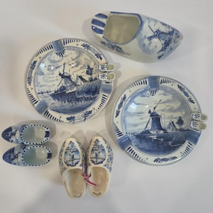 May include: A collection of blue and white ceramic items, including ashtrays with windmill and ship designs, and clogs. The ashtrays are round with a rim design. The clogs are in pairs, one large and two small.