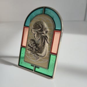 May include: A silver-toned religious icon featuring a mother and child, framed by a stained-glass effect with teal, pink, and green accents. The artwork is set against a gold background with etched lines, creating a vintage aesthetic.