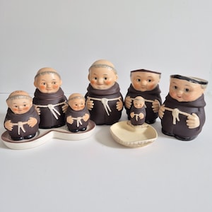 May include: A collection of ceramic monk figurines in brown robes with white rope ties. The set includes salt and pepper shakers, a small dish, and a larger container. The figurines have smiling faces and are arranged on a white surface.