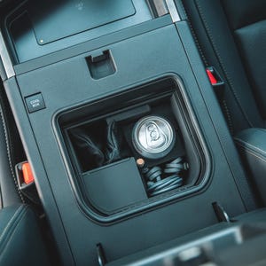 May include: A car's center console with a built-in cooler. Inside, a silver can of soda, a small black box, and a bundle of black cables are visible. The console is black, with a button labeled "COOL BOX". The surrounding seats are black leather.