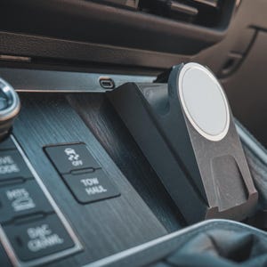 以下が含まれることがあります： 車の内装のクローズアップ。黒いダッシュボードに木目調のアクセント。ドライブモードセレクター、オフロードと牽引機能のボタン、白い円形パッド付きの電話マウントが含まれています。
