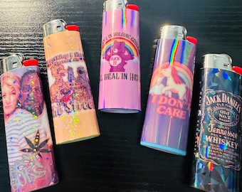 May include: Five iridescent lighters with various designs. One features a rainbow and a unicorn with the text "I DON'T CARE". Another has a bear with a rainbow and the text "FUCK IT". Other lighters have images of celebrities and cartoon characters.