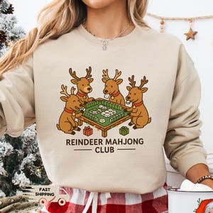 Puede incluir: Sudadera color arena con un gr&aacute;fico de cuatro renos jugando al mahjong. El texto "REINDEER MAHJONG CLUB" est&aacute; debajo de la ilustraci&oacute;n. Parte de una colecci&oacute;n tem&aacute;tica navide&ntilde;a.