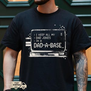 Könnte beinhalten: Schwarzes T-Shirt mit einem weißen Grafikdesign eines Retro-Computerbildschirms mit dem Text "I KEEP ALL MY DAD JOKES IN A DAD-A-BASE_". Mit "Schneller Versand" Logo.