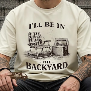 May include: Ivory-colored t-shirt with a graphic that reads "I'LL BE IN THE BACKYARD." The graphic depicts a folding chair, a cooler, a baseball cap, and a blanket. The shirt is a relaxed fit.